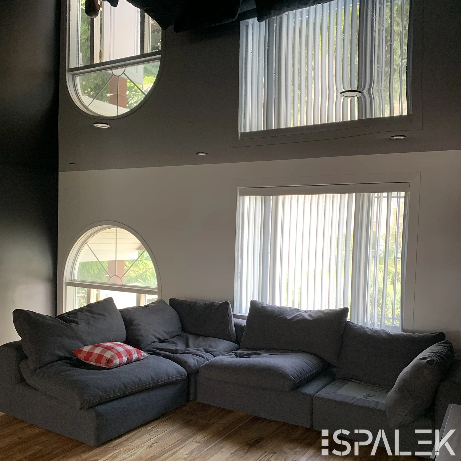 Modernes Wohnzimmer mit grauem Ecksofa und rot-kariertem Kissen vor weißer Wand, großen Fenstern, Vertikaljalousien und Lichtleiste.