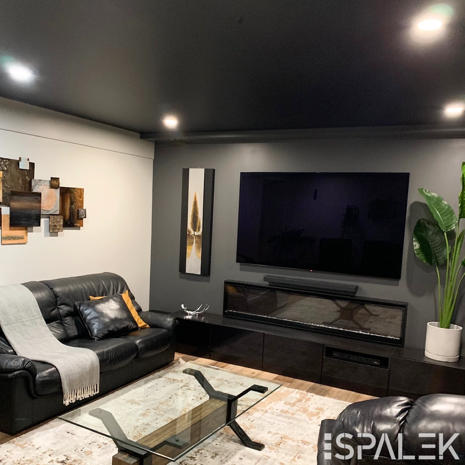 Modernes Wohnzimmer mit schwarzen Ledersofas, Glastisch, TV über Elektrokamin, abstrakter Wandkunst und eleganter Lichtleiste.