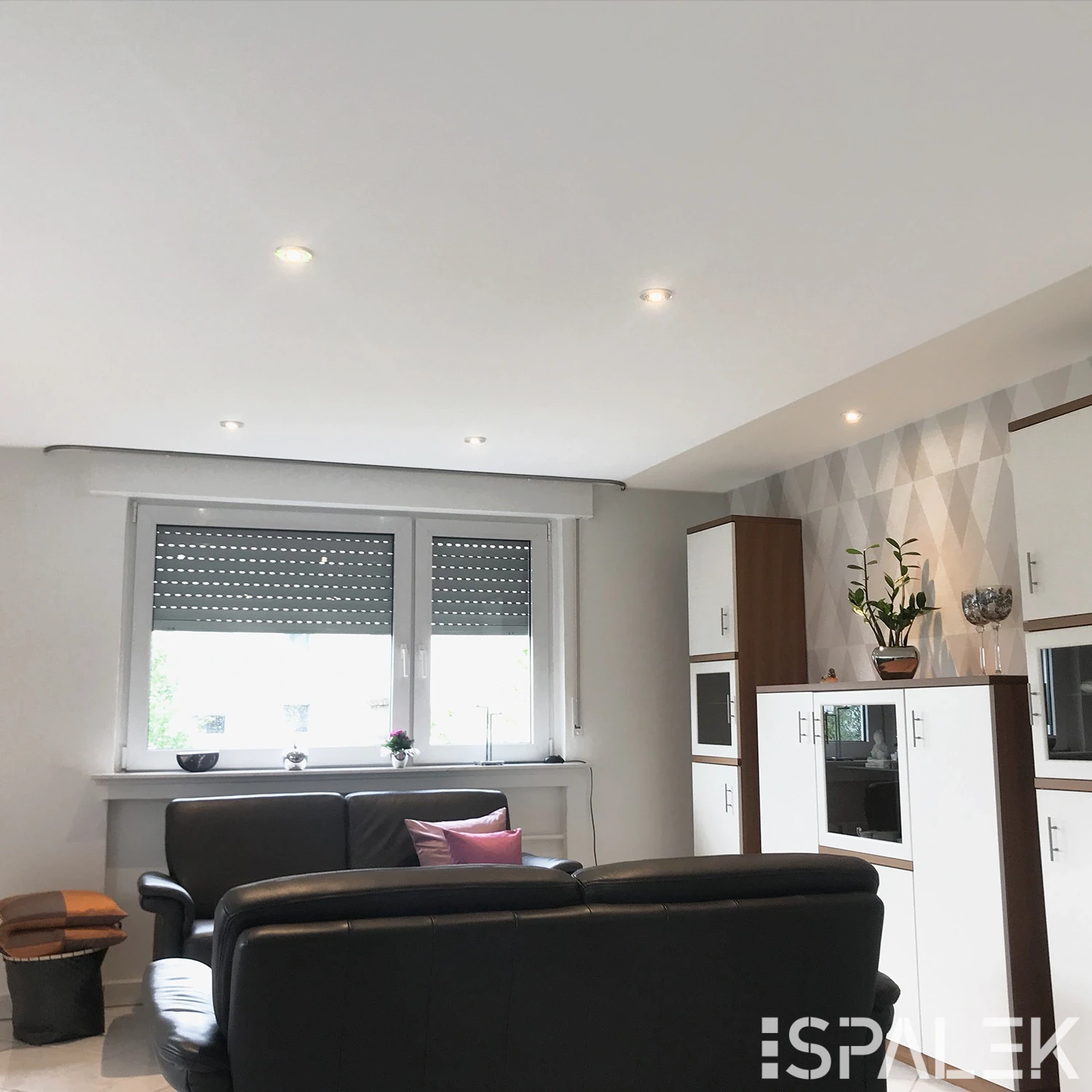 Modernes Wohnzimmer mit zwei schwarzen Ledersofas, Fenster mit geschlossenen Jalousien, weißen Schränken und Pflanzen.