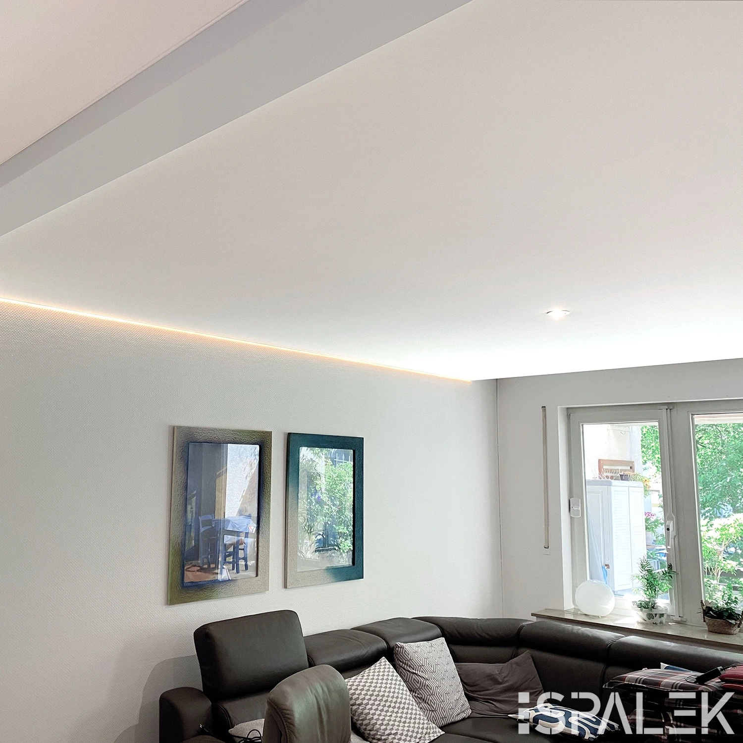 Modernes Wohnzimmer mit grauem Ecksofa, gemusterten Kissen, zwei gerahmten Bildern und großen Fenstern. LED-Lichtleiste an der Decke.
