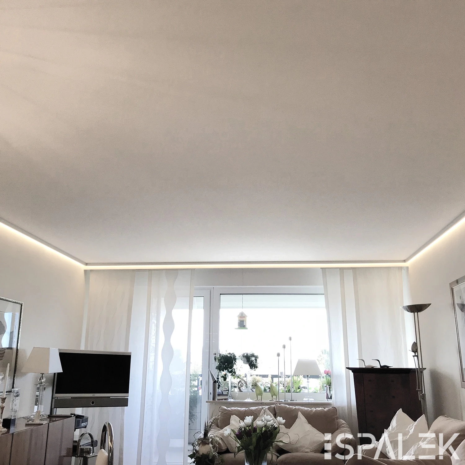 Helles Wohnzimmer mit großen Fenstern, weißen Gardinen, beigen Sofas, TV, Lampen, Pflanzen und indirekter Lichtleiste an der Decke.