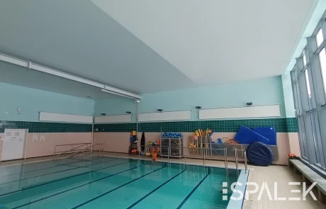 Hallenbad mit klarem Wasser, Metallhandläufen und großen Fenstern rechts. Bunte Poolgeräte an der grün gefliesten Wand.
