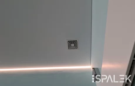 Die Decke im DRK Schwimmbad Raisdorf zeigt mittig ein quadratisches Lüftungsgitter und einen schmalen LED-Lichtstreifen am Rand. In einer Ecke sind Teile einer Lüftungsanlage sowie eine weiße Wand erkennbar.