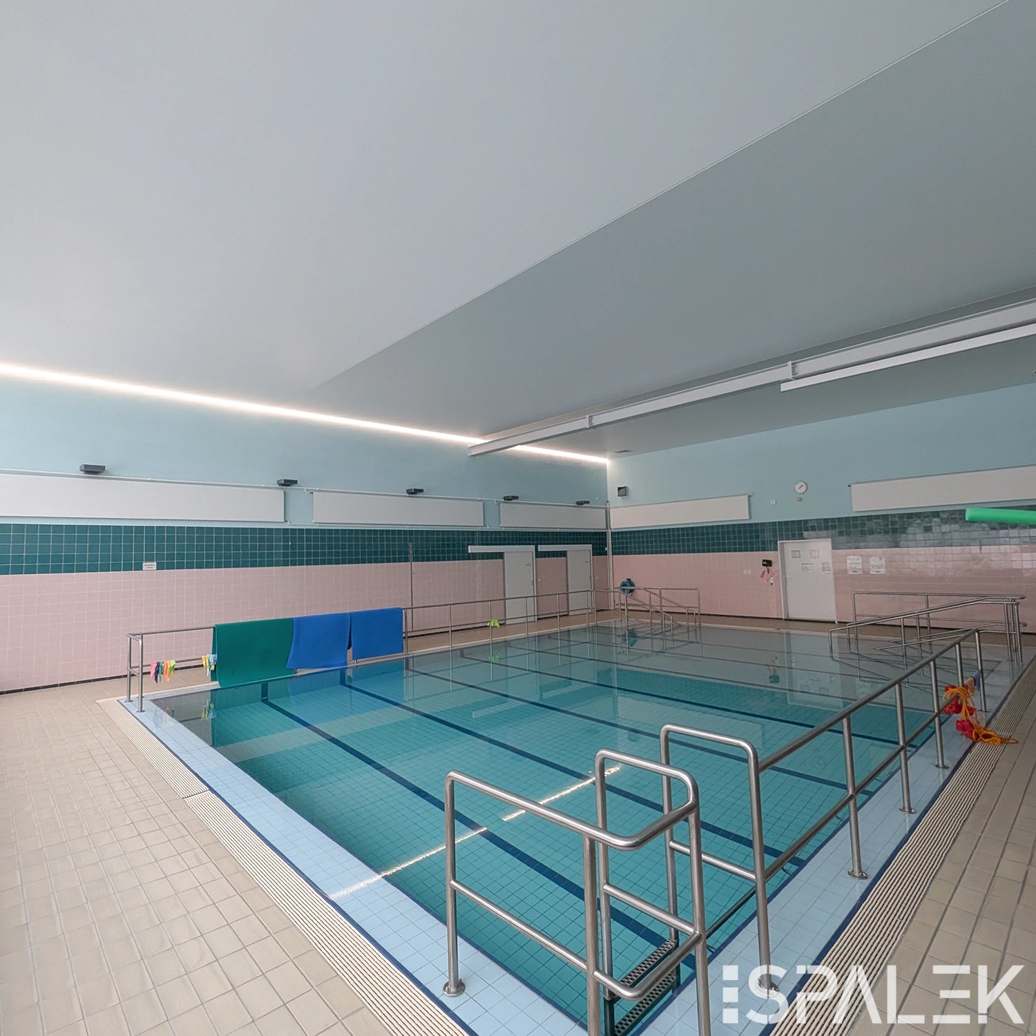 Das Hallenbad DRK Schwimmbad Raisdorf mit blauem Wasser, Metallgeländern und Fliesen. Die Halle ist gut beleuchtet.