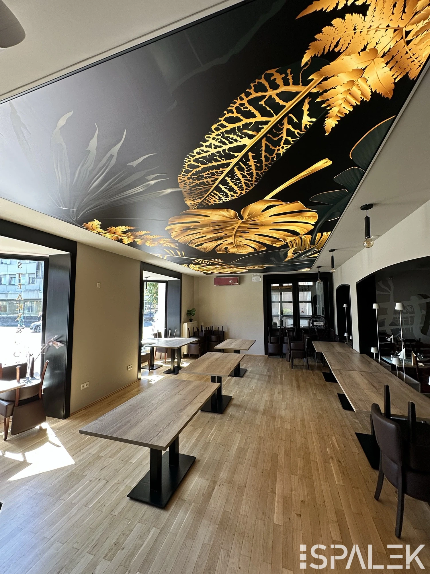 Hotel Heinsberg Modernes Restaurant mit Holzboden, großen Fenstern und leeren Tischen. Die Decke zeigt ein auffälliges Blattmuster in Goldgelb auf Schwarz.