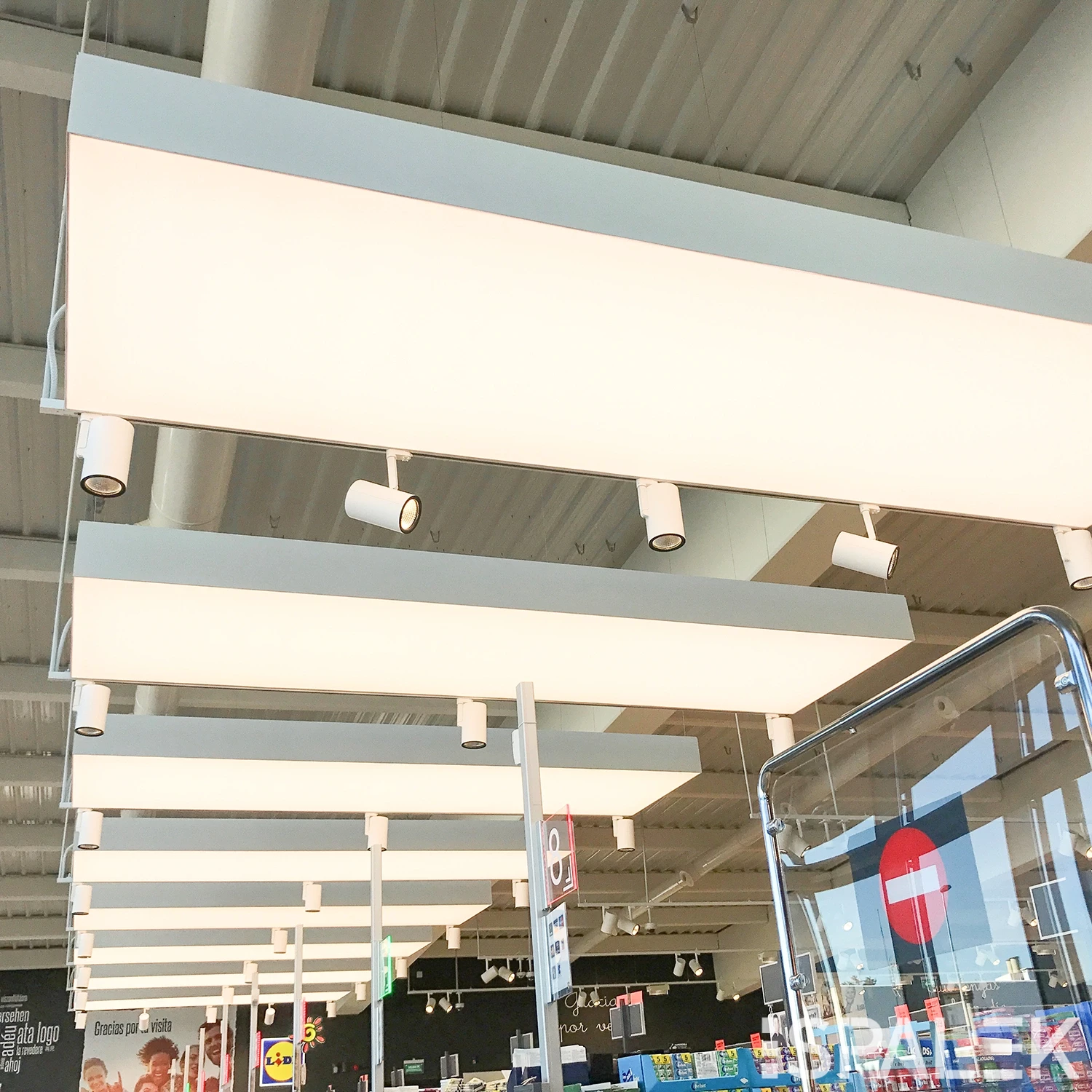 image001 Helle Deckenleuchten und Spotlights hängen von einer weißen, industriellen Decke über einem modernen Supermarkt mit Glasabtrennung und sichtbaren Schildern.