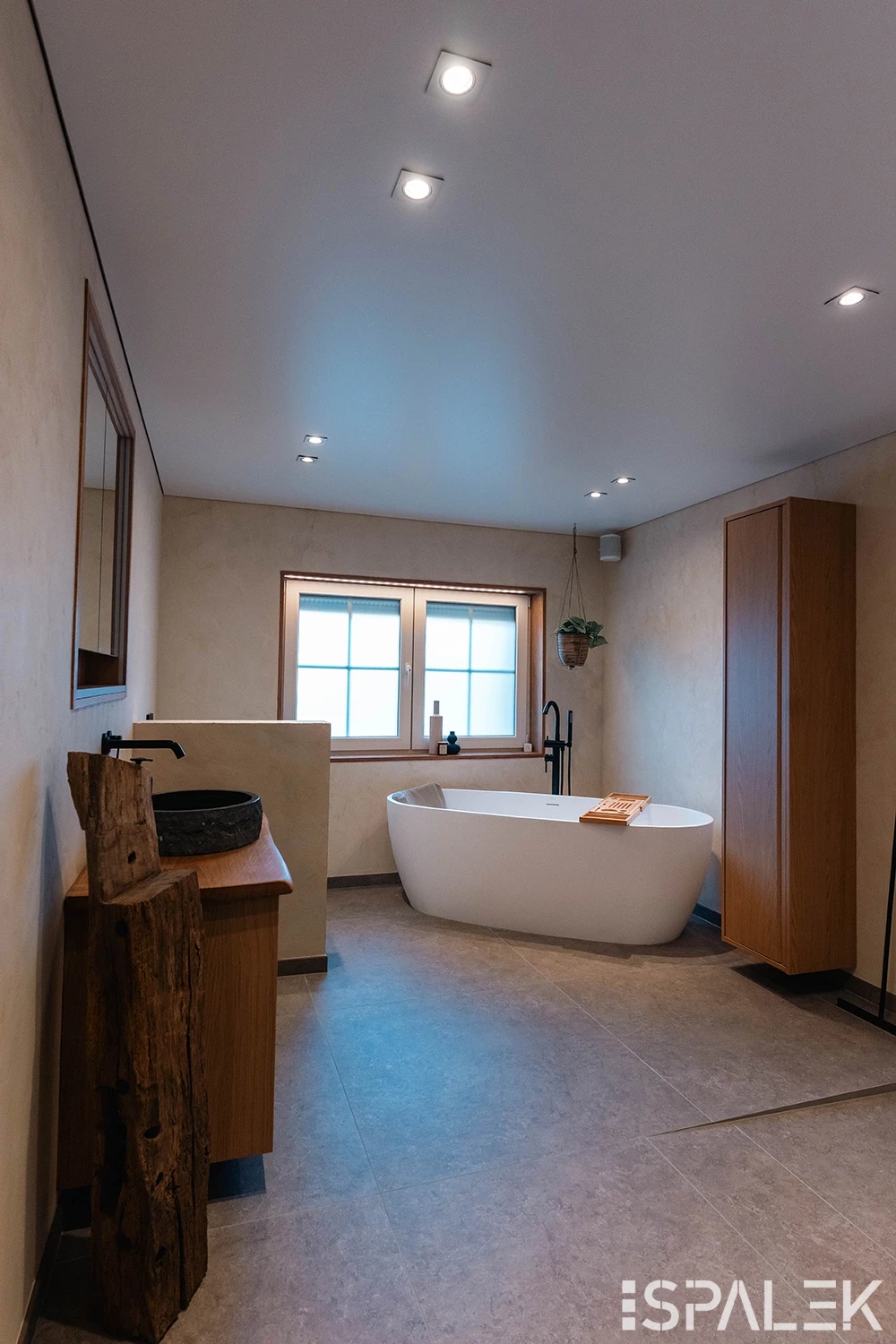 Modernes Bad mit freistehender weißer Badewanne, Holzmöbeln, Aufsatzwaschbecken, viel Tageslicht und grünen Zimmerpflanzen.