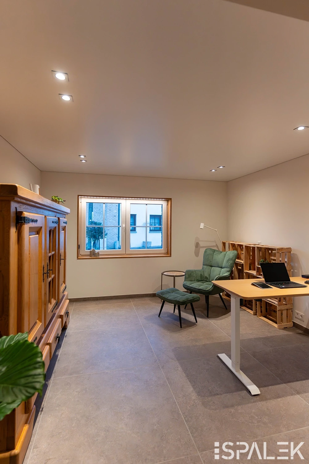 Minimalistisches Homeoffice mit Holzschrank, grünem Sessel und Hocker, kleinem Tisch, Regal, Laptop auf weißem Schreibtisch und Fenster.