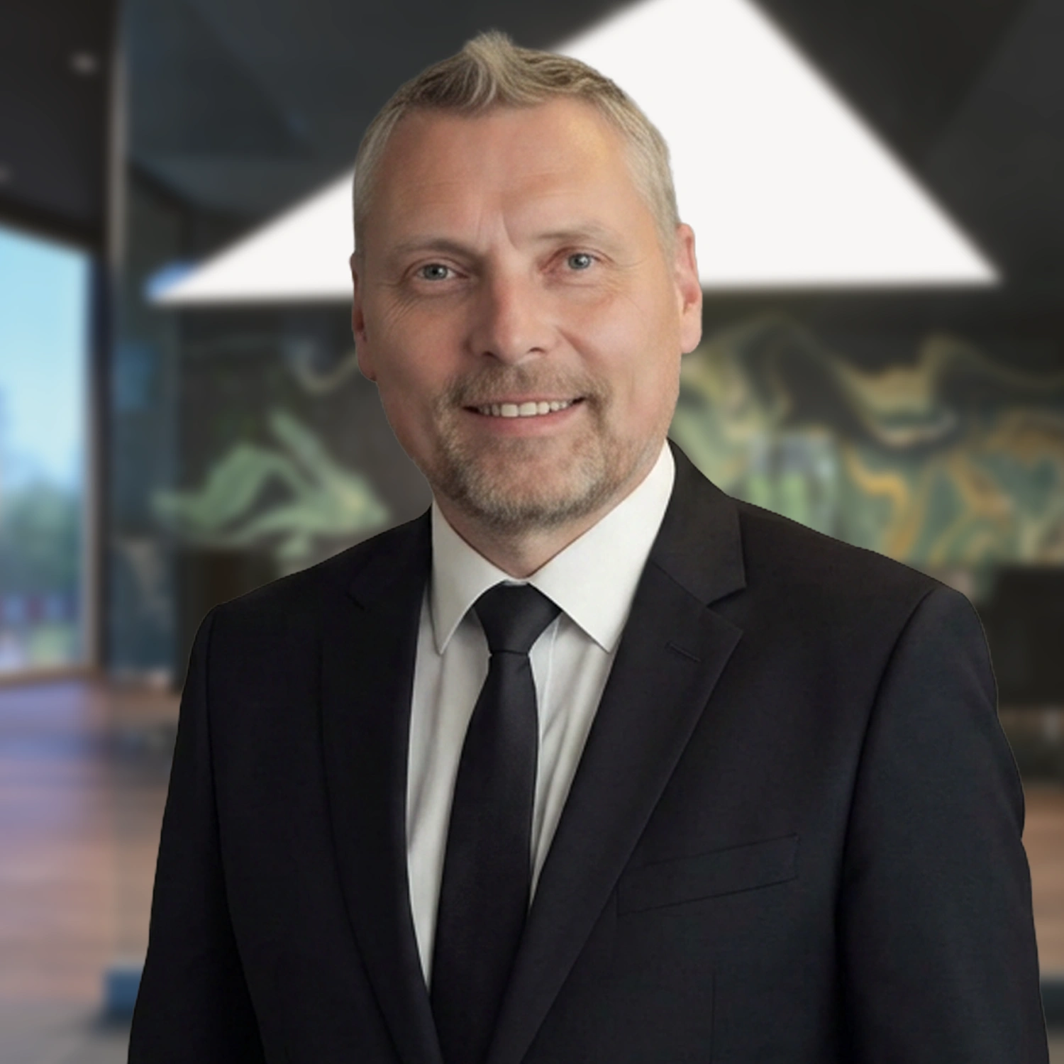Ein mittelalterlicher Mann mit blondem Kurzhaarschnitt und Bart trägt Anzug und Krawatte und steht lächelnd in einem modernen Büro mit abstrakter Wandkunst und großen Fenstern.