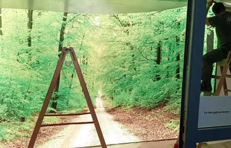 Ein Mitarbeiter bringt ein Waldmotiv an einer Wand an. Eine Leiterin steht daneben, "Mitten im Wald" und "SPA LEK" sind zu sehen.