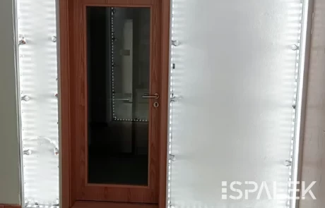 Holztür mit Glaseinsatz in weißer Wand, umrahmt von sichtbaren LED-Streifen. Grünes und beiges Bodendesign, Kabel hängt ab.