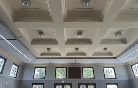 Decke eines Raums im Bahnhof Wittenberge mit quadratischen, eingelassenen Feldern, in denen jeweils ein kleiner weißer Lautsprecher oder Sensor angebracht ist. An den oberen Wänden befinden sich große rechteckige Fenster.