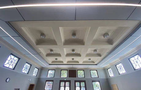 Deckenansicht eines modernen Raums mit eingelassenen quadratischen Paneelen, mehreren Deckenleuchten und großen Fenstern mit geometrischem Muster, durch die Tageslicht hereinfällt. An einer Wand befinden sich eine Uhr und zweiflügelige Holztüren.