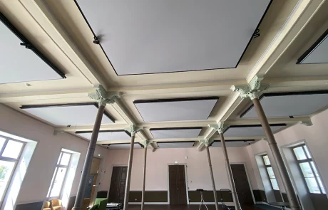 Ein großzügiger Raum mit hoher Decke, quadratischen Akustikpaneelen, Säulen, großen Fenstern und Türen. Entlang der Wände stehen Sofas, ein Stuhl und ein kleiner Tisch; Sonnenlicht fällt herein.