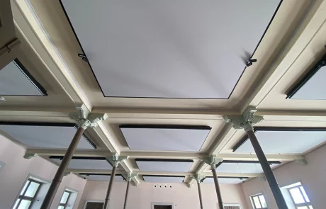 An der Decke eines großen Raums mit Säulen und dekorativen Kapitellen sind rechteckige Akustikpaneele angebracht. Die Wände sind beige, es gibt mehrere Fenster und Türen.