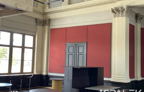 Fertig1 Ein großzügiger Raum mit hohen Fenstern, roten Wandpaneelen, dekorativen Säulen und übereinander gestapelten Möbeln. Sonnenlicht fällt durch die Fenster und wirft schattierte Muster auf den Boden. Am rechten unteren Bildrand ist der Schriftzug „SPAŁEK“ zu sehen.