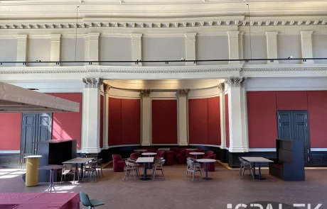 Fertig2 Geräumiger Raum mit hoher Decke, roten Wandpaneelen und klassischen Säulen. Mehrere Tische und Stühle sind im Raum verteilt angeordnet. Natürliches Licht fällt von außen herein, unten rechts ist das ESPALEK-Logo zu sehen.