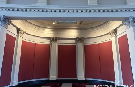 Fertig3 Eine geschwungene Wand mit roten Paneelen und verzierten weißen Säulen umrahmt einen Sitzbereich mit roten Stühlen und runden Tischen unter einer hohen Decke. Der Raum ist leer und hell beleuchtet.