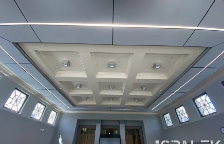 Seminar24fertig2 Moderne Kassettendecke mit geometrisch angeordneten, eingelassenen Leuchten, umgeben von Oberlichtern mit geometrischen Sprossen; der ruhige Raum wirkt wie mitten im Wald, was durch eine neutrale Farbgestaltung und symmetrische architektonische Details noch verstärkt wird.