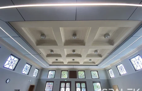 Seminar24fertig6 Ein großer, moderner Raum mit Kassettendecke und eingelassenen Leuchten. Hohe Fenster mit geometrischen Mustern lassen Tageslicht herein und erhellen den Raum. An der linken Wand hängt eine Uhr, am gegenüberliegenden Ende befinden sich doppelte Holztüren.