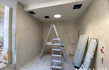SepareeimBau Baustelle mit beigen Wänden, einer Aluminiumleiter in der Mitte, Schalttafeln an der rechten Wand und Deckenöffnungen. Der unfertige Boden und ein Oberlicht sorgen für eine offene Atmosphäre in diesem abgeschiedenen Innenraum.