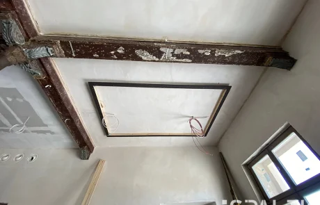 Stützensaalvorher2 Ein renovierungsbedürftiger Raum mit freiliegenden Holzbalken, einem quadratischen Deckenrahmen und herabhängenden Kabeln, unverputzten Wänden und einem kleinen Fenster auf der rechten Seite. Im rechten unteren Bildbereich ist das ESPALEX-Logo zu sehen.
