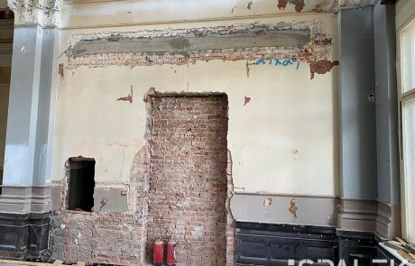 vorher2 Freigelegte Backsteinwand mit zwei zugemauerten Türöffnungen und teilweise abgeplatztem Putz in einem alten, teilweise renovierten Gebäude. Zwei rote Feuerlöscher stehen vorne auf dem Boden und setzen einen modernen Akzent in der rustikalen Umgebung.